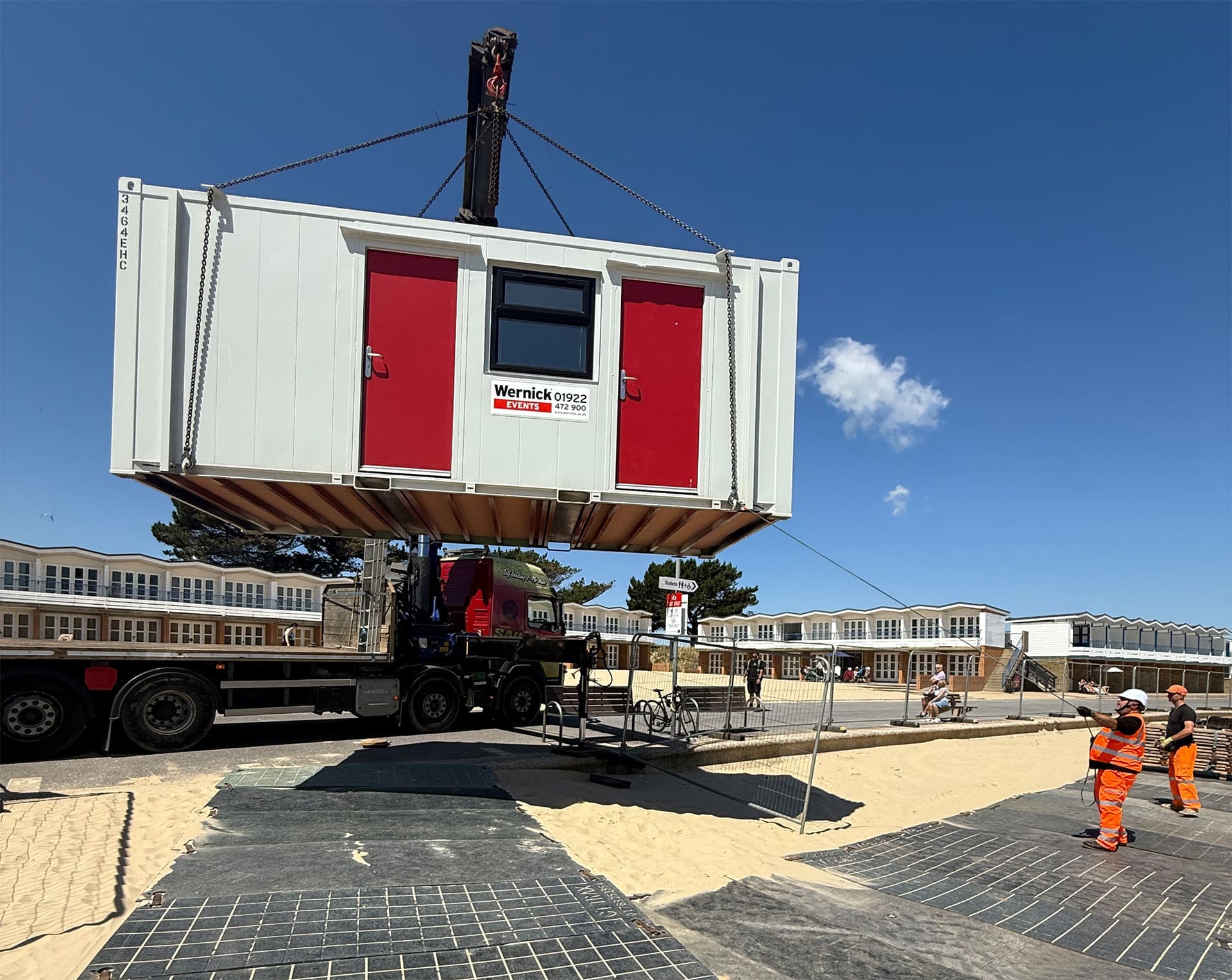 A crane lorry carefully moving a production office on to sire for a festical build on the beach, with staff carefully using the correct PPE